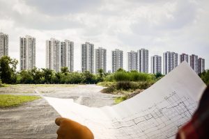 Profissional segura planta arquitetônica diante de um grande terreno aberto, com prédios altos ao fundo, representando planejamento urbano e desenvolvimento imobiliário.