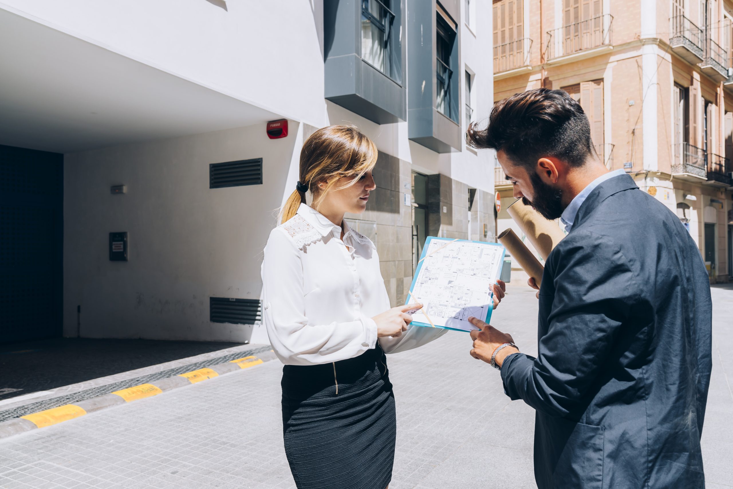 Mulher e homem analisando uma planta de imóvel em uma rua urbana, ao lado de um edifício moderno.