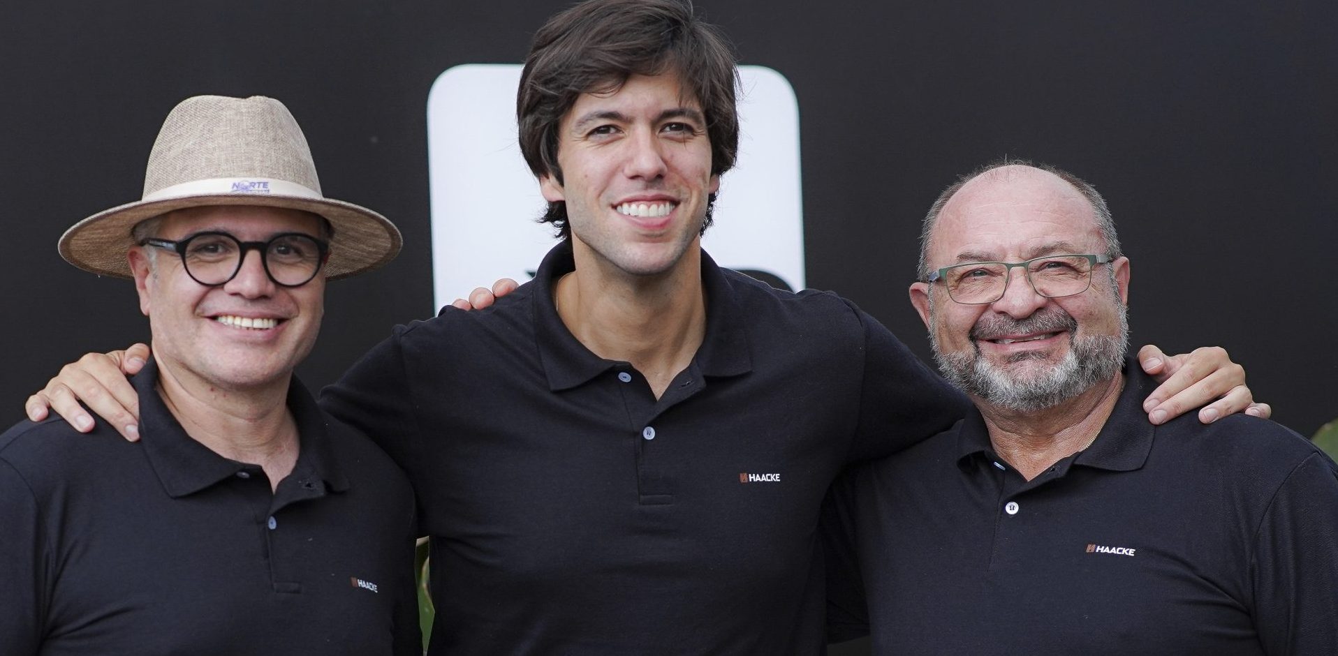 Três homens sorridentes usando camisetas pretas com o logo “HAACKE”, posando juntos com os braços nos ombros uns dos outros.