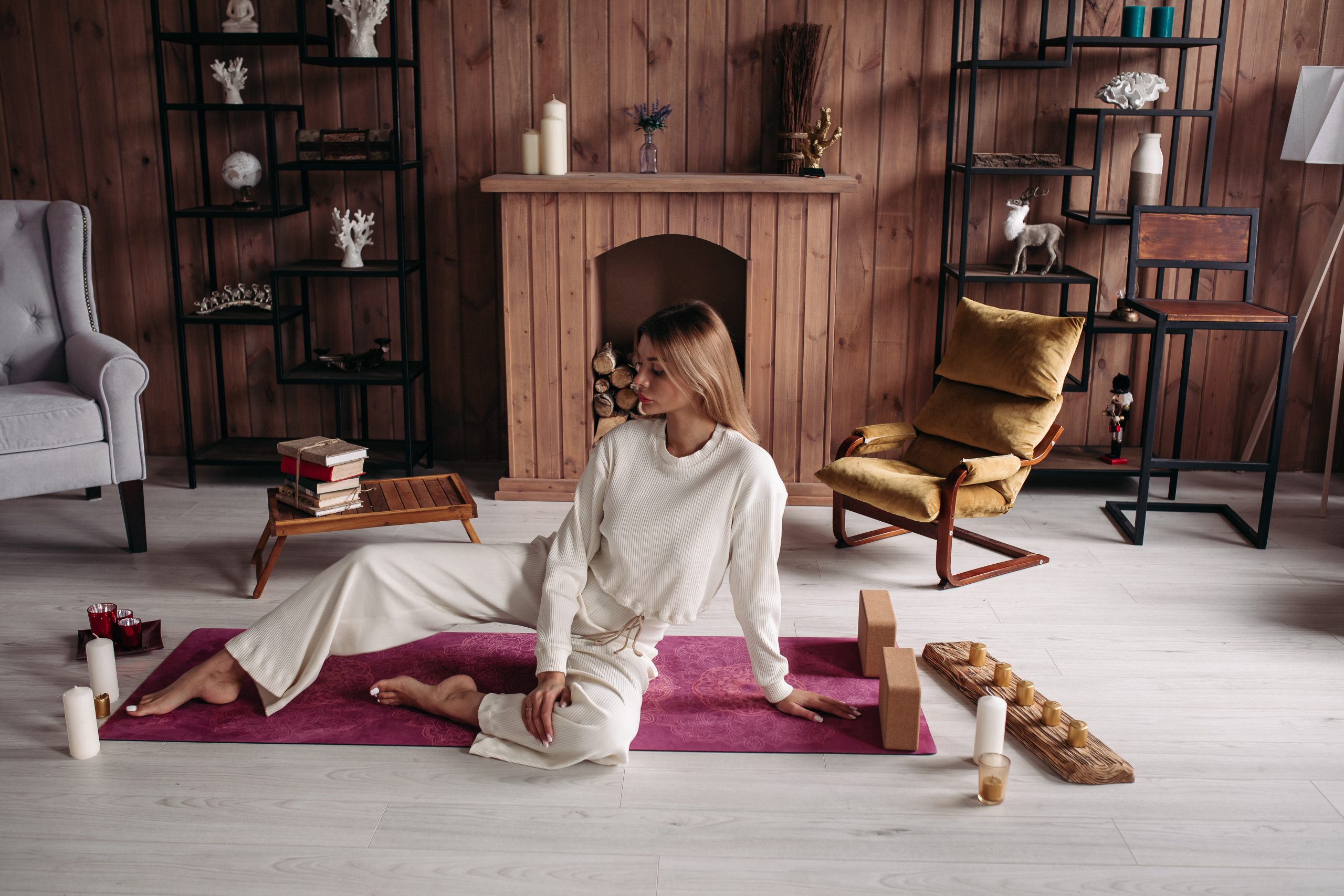 Mulher sentada em um tapete de yoga em um ambiente aconchegante com decoração rústica e elementos de bem-estar ao redor.