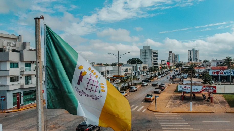 Bandeira do município de Primavera do Leste tremulando em um mastro com vista para uma avenida central da cidade.