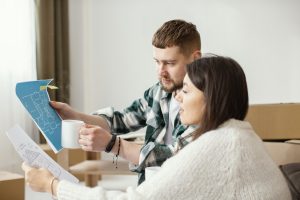 Casal analisando plantas de imóvel enquanto seguram xícaras de café, em ambiente com caixas de mudança.