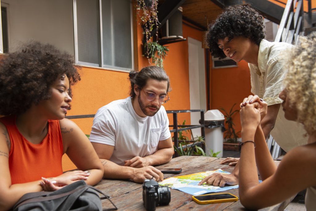 Grupo de quatro jovens observando um mapa sobre a mesa, planejando uma viagem em ambiente descontraído com paredes laranja ao fundo.