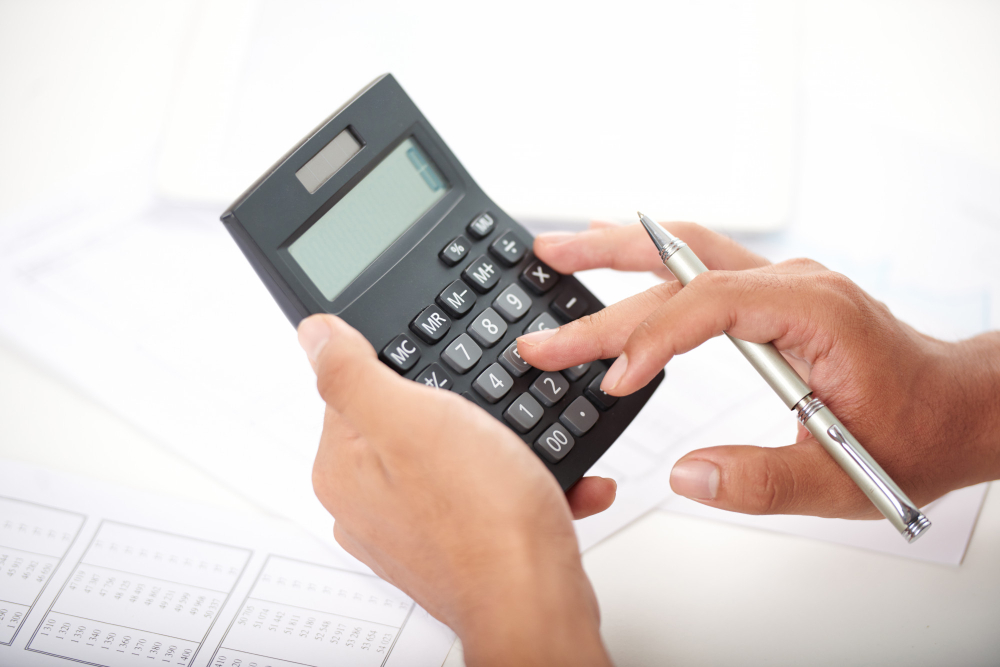 Pessoa segurando uma calculadora e digitando números com uma mão, enquanto segura uma caneta com a outra, sobre uma mesa com papéis.