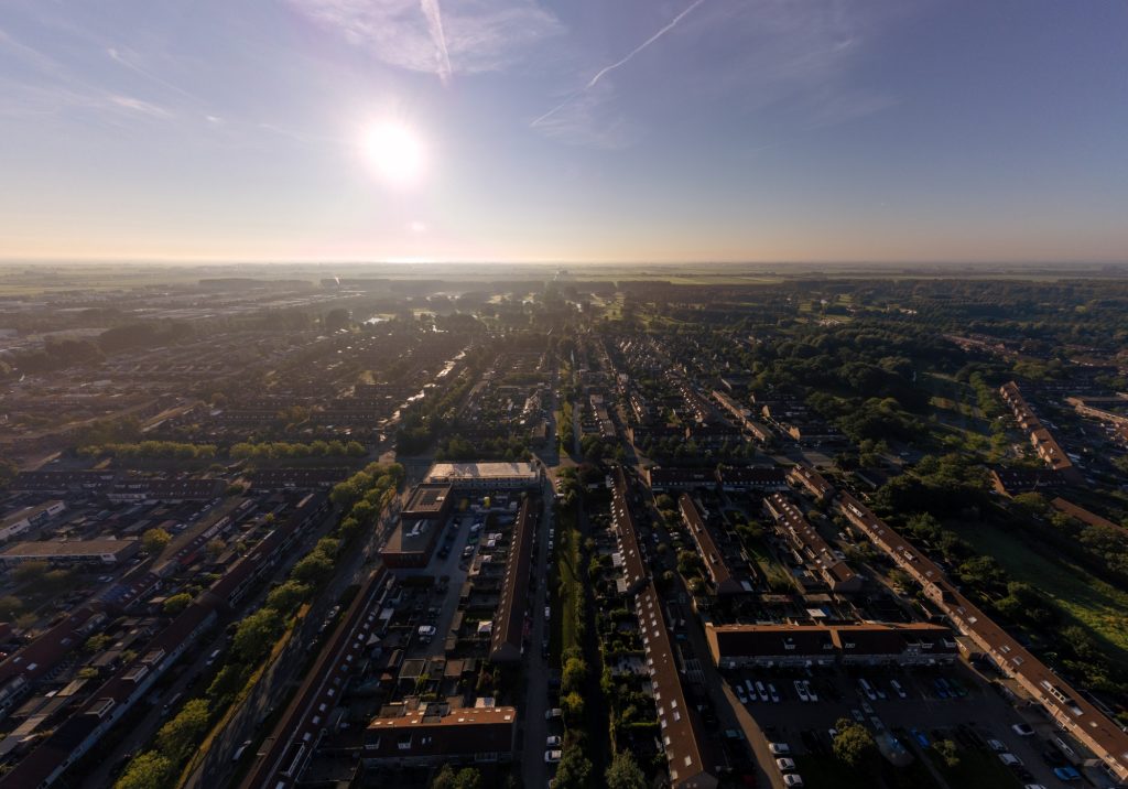 Vista aérea de uma cidade ao entardecer, com o sol brilhando forte no horizonte e projetando longas sombras sobre os prédios e ruas alinhadas. A paisagem urbana é cercada por áreas verdes e campos ao fundo.