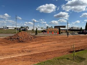 Área de construção com solo avermelhado, entulho e cones de sinalização, sob céu azul com nuvens.