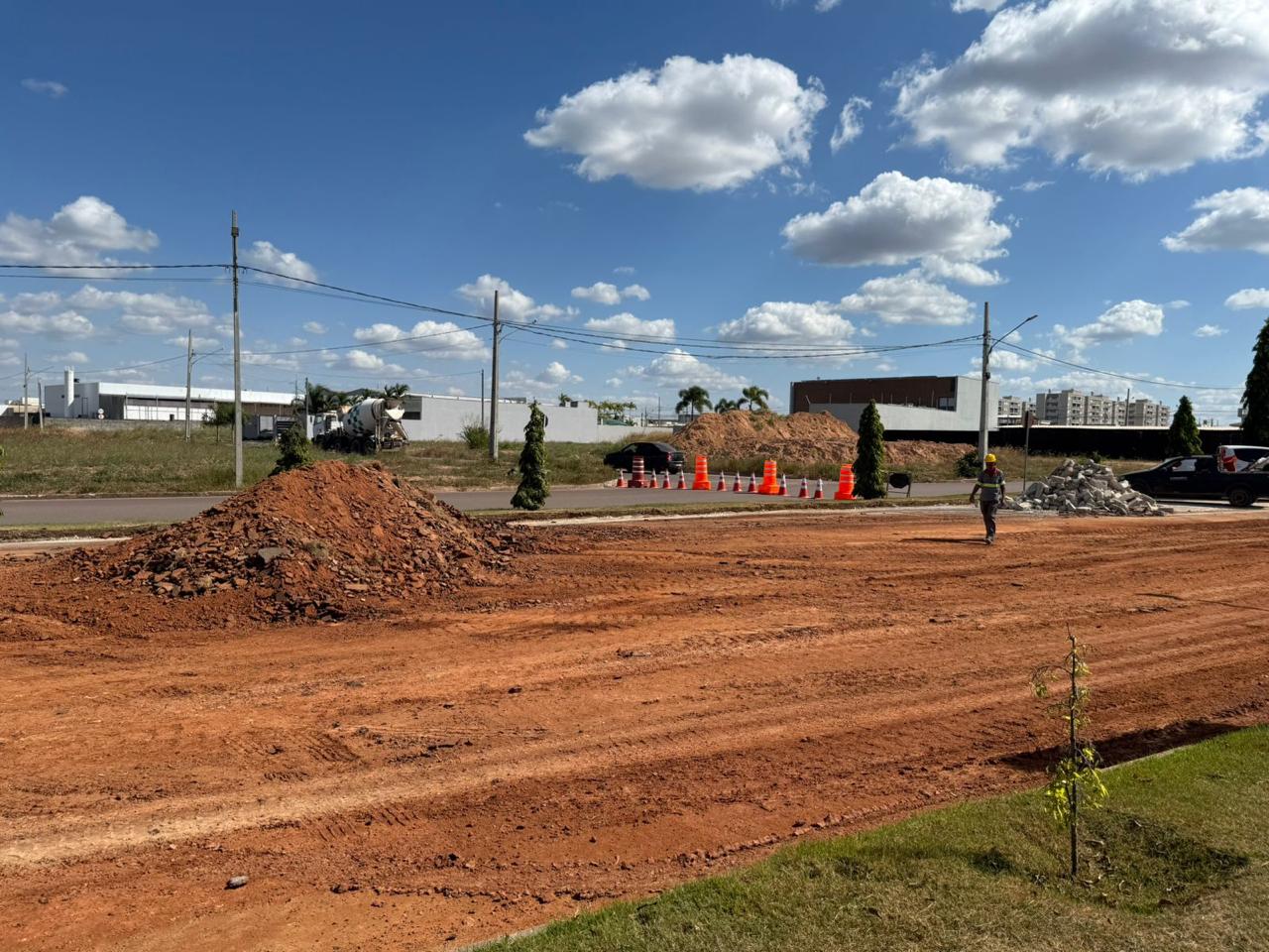 Área de construção com solo avermelhado, entulho e cones de sinalização, sob céu azul com nuvens.