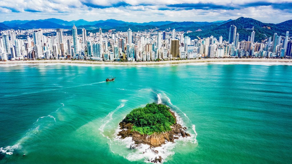 Vista aérea de uma pequena ilha com vegetação cercada por mar verde, com a cidade de Balneário Camboriú ao fundo.