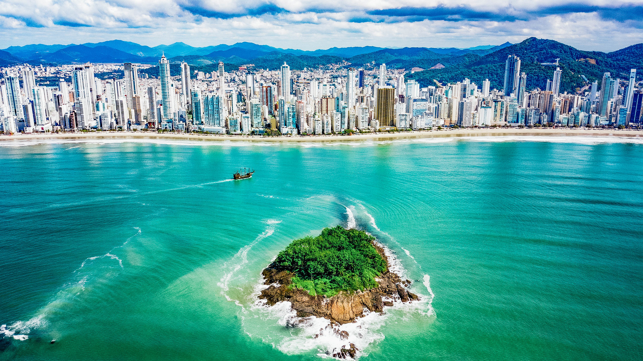 Vista aérea de uma pequena ilha com vegetação cercada por mar verde, com a cidade de Balneário Camboriú ao fundo.