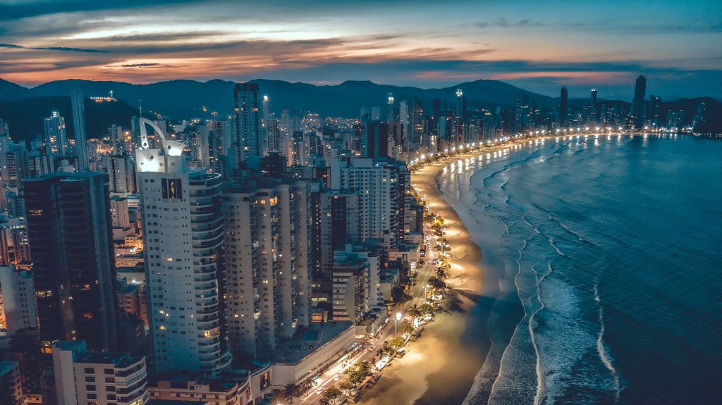 Vista aérea noturna da orla de Balneário Camboriú iluminada, com arranha-céus e mar calmo ao entardecer.
