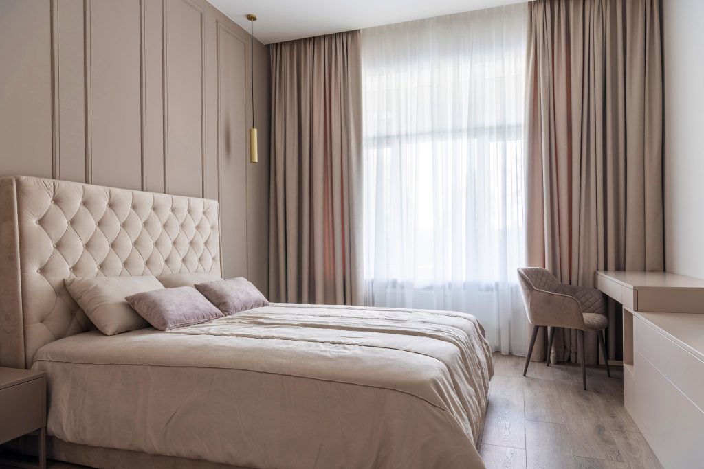 Quarto moderno com cama de casal bege, cabeceira acolchoada, cortinas longas e escrivaninha com cadeira estofada ao lado da janela.