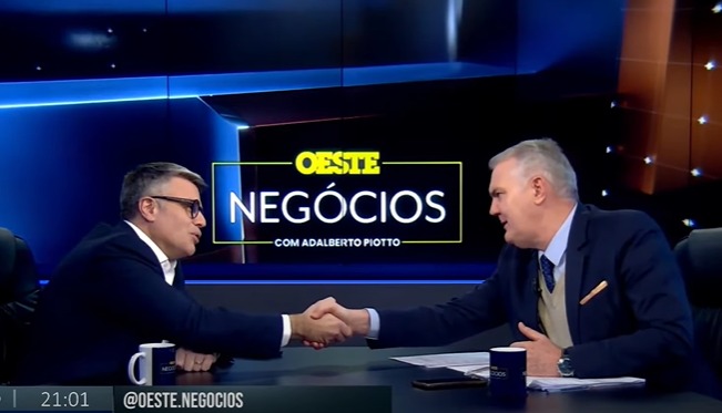 Dois homens de terno se cumprimentam com um aperto de mãos durante o programa "Oeste Negócios", apresentado por Adalberto Piotto.