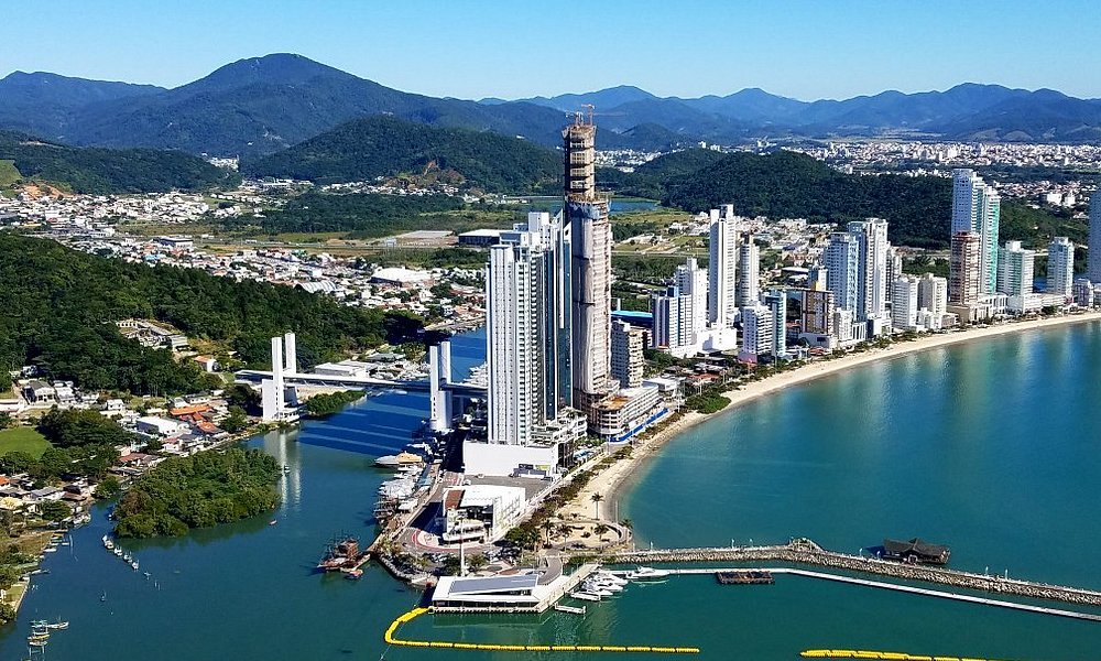 Vista aérea de Balneário Camboriú, em Santa Catarina, com arranha-céus à beira-mar, mar azul esverdeado e montanhas ao fundo.