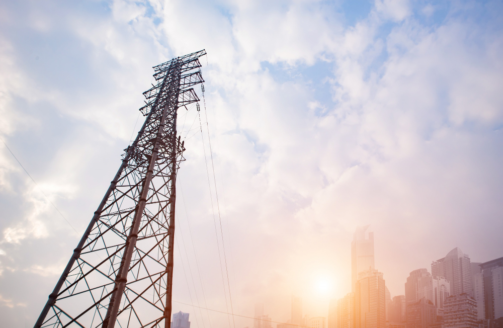 Torre de transmissão elétrica em frente a prédios urbanos com o sol nascendo, simbolizando energia e conectividade digital.