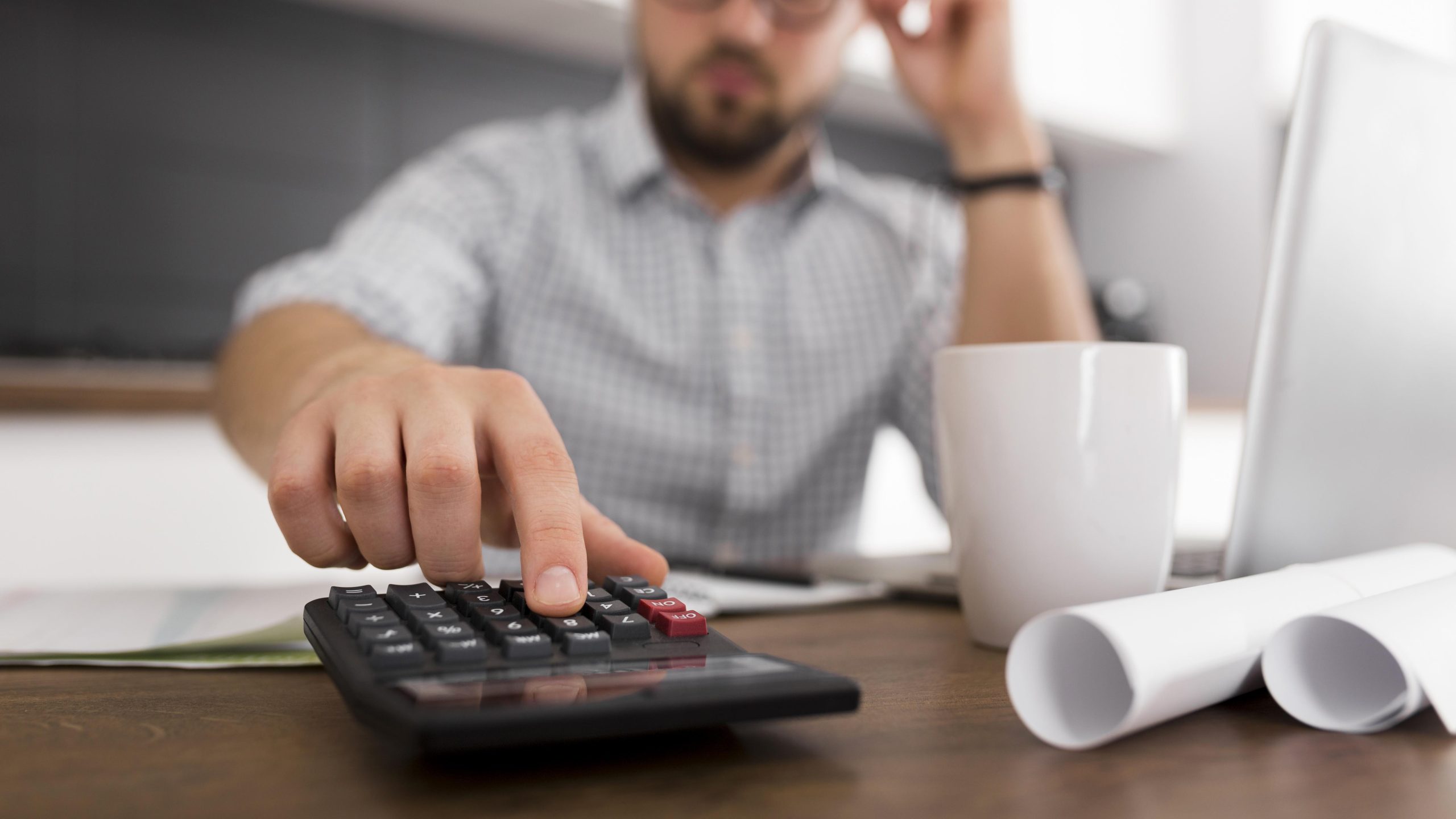 Homem usando uma calculadora sobre uma mesa de escritório, com caneca branca, documentos enrolados e notebook ao fundo.