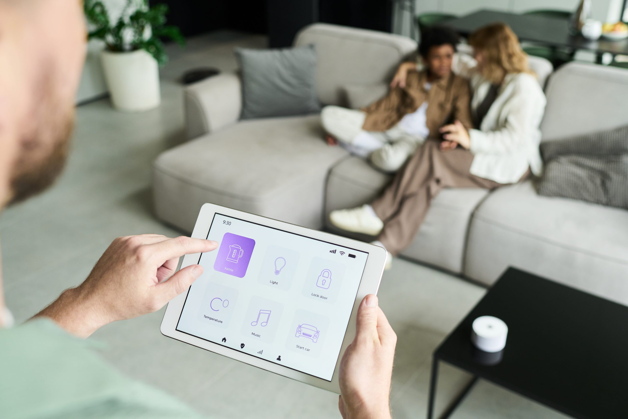 Mãos segurando um tablet com aplicativo de casa inteligente, com um casal relaxando no sofá ao fundo.
