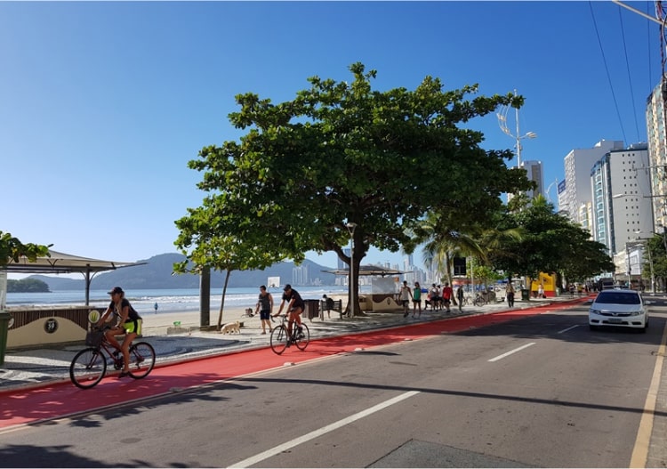 A imagem mostra uma avenida litorânea arborizada, com uma ciclovia vermelha paralela à calçada da praia. Algumas pessoas pedalam de bicicleta e outras caminham próximo à areia.