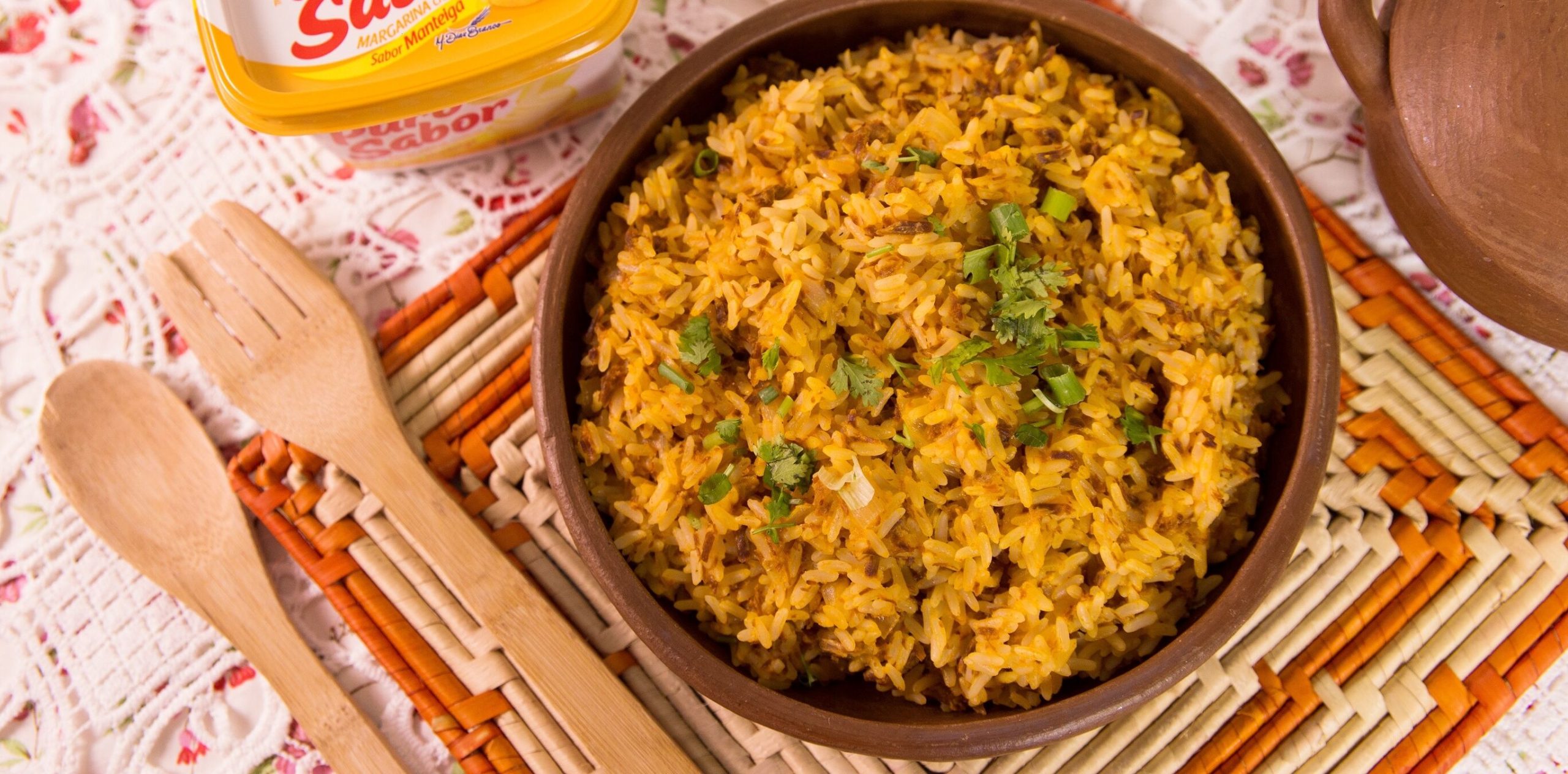 Prato de Arroz Maria Isabel em uma tigela de madeira, com duas colheres de madeira ao lado, sobre uma toalha de mesa e um pote de manteiga ao fundo.