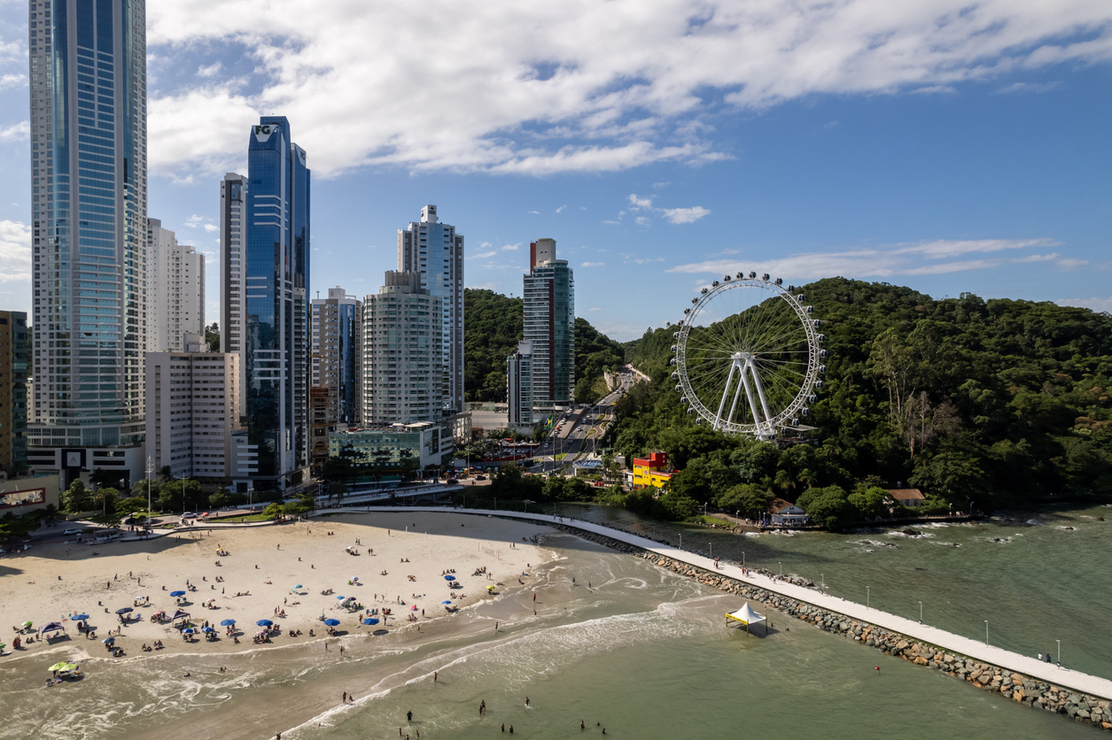 Vista aérea de Balneário Camboriú com a praia, o molhe, os prédios altos e a roda-gigante FG Big Wheel ao fundo, cercada pela Mata Atlântica.