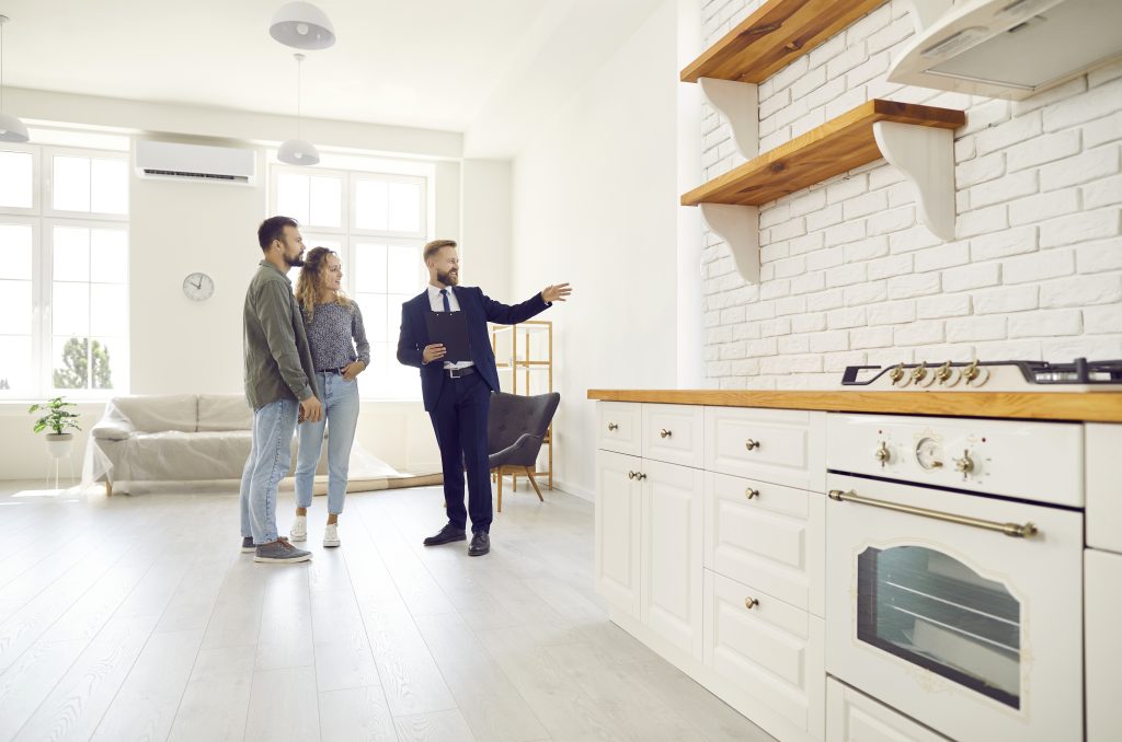 Corretor de imóveis apresentando uma cozinha moderna de apartamento para um casal jovem, com armários brancos e bancada de madeira em ambiente iluminado.