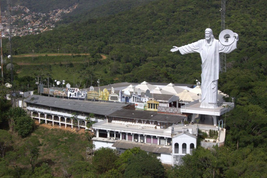 Estátua gigante de Jesus Cristo em meio a uma área verde, com construções religiosas ao redor.