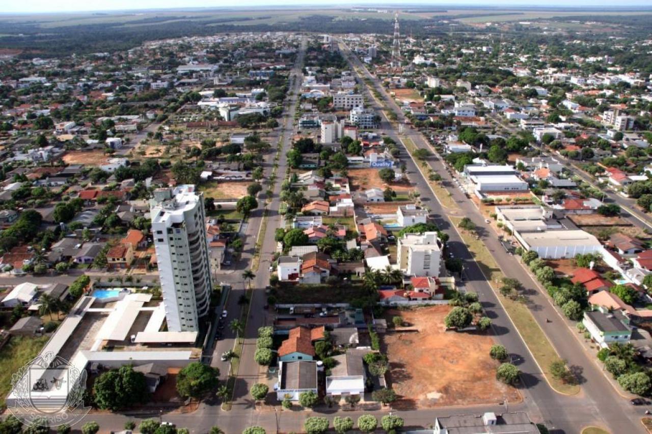 Vista aérea de Sorriso, Mato Grosso, mostrando a área urbana da cidade. A imagem destaca uma avenida principal que corta a cidade, cercada por construções baixas, algumas árvores e lotes de terra vazios.