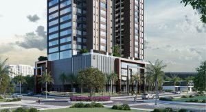 Renderização realista de um empreendimento residencial de alto padrão com duas torres modernas, fachada em tons de marrom e cinza, grandes janelas espelhadas e áreas verdes no topo. A base do edifício apresenta um design contemporâneo com linhas verticais e detalhes em vidro. Em primeiro plano, há uma rotatória com canteiro florido, árvores, palmeiras e pessoas caminhando, transmitindo um ambiente urbano sofisticado e bem planejado.
