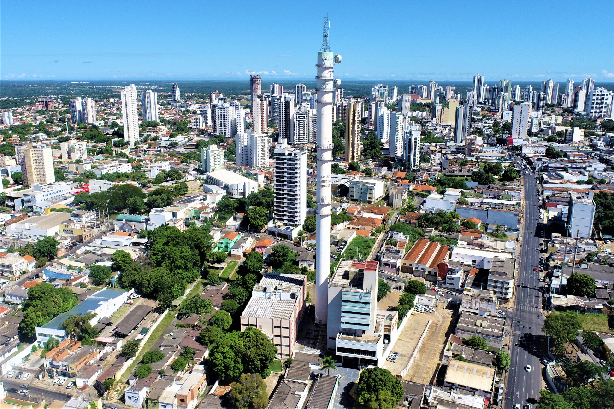 Vista aérea de uma cidade com muitos prédios altos, áreas residenciais e uma torre de telecomunicações no centro da imagem.