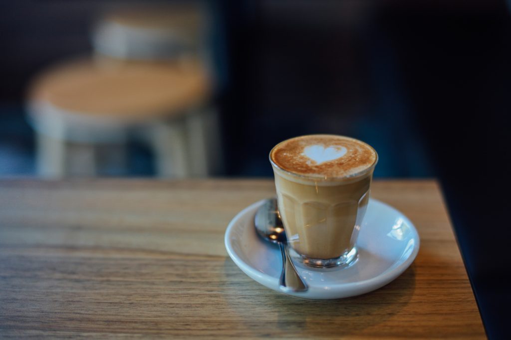 Xícara de café com leite em copo de vidro, com desenho de coração na espuma, sobre mesa de madeira.