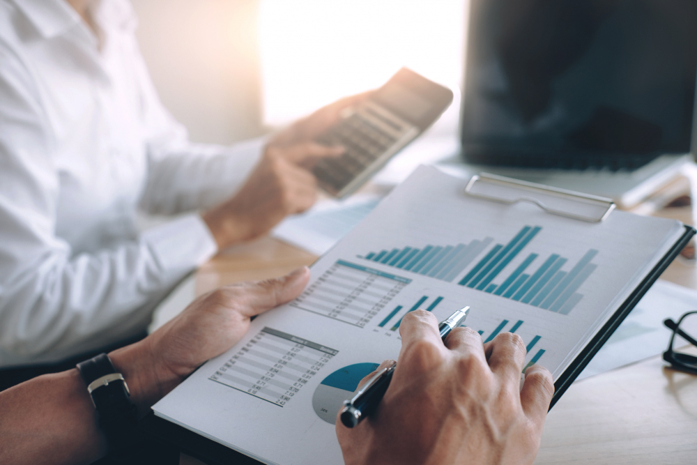 Pessoa analisando gráficos financeiros em prancheta, com calculadora na mão, em ambiente corporativo.