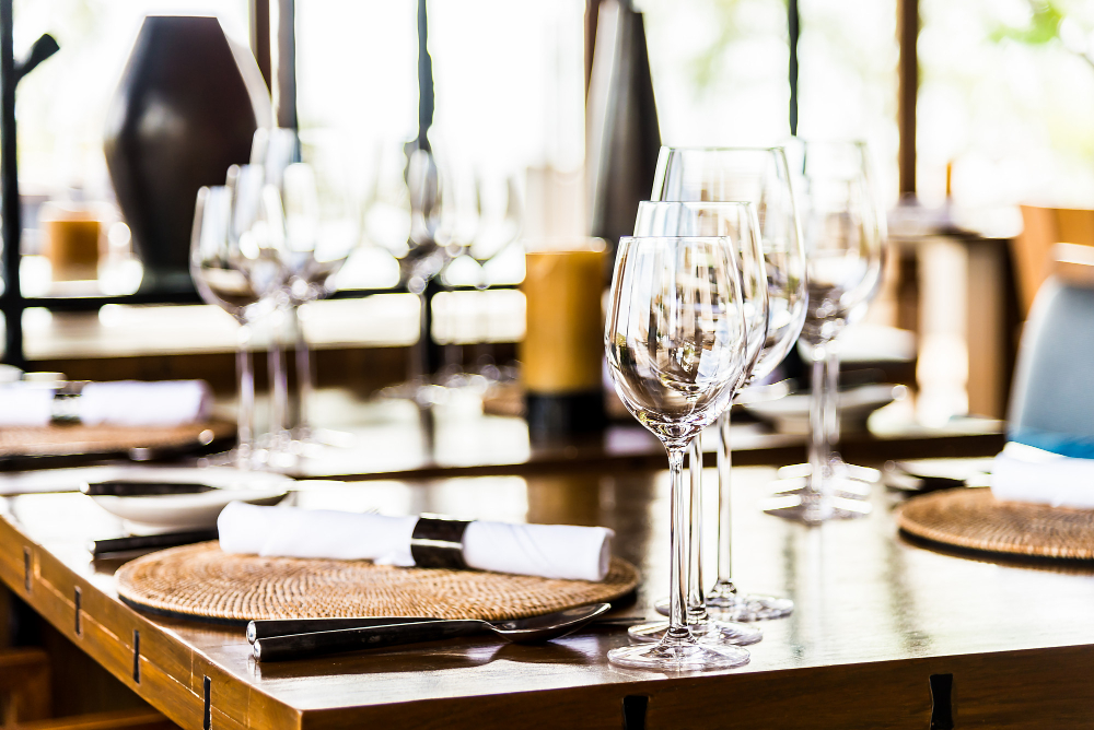 Mesa de restaurante elegante com taças de vinho alinhadas, pratos com sousplats e talheres organizados, em ambiente sofisticado e bem iluminado.