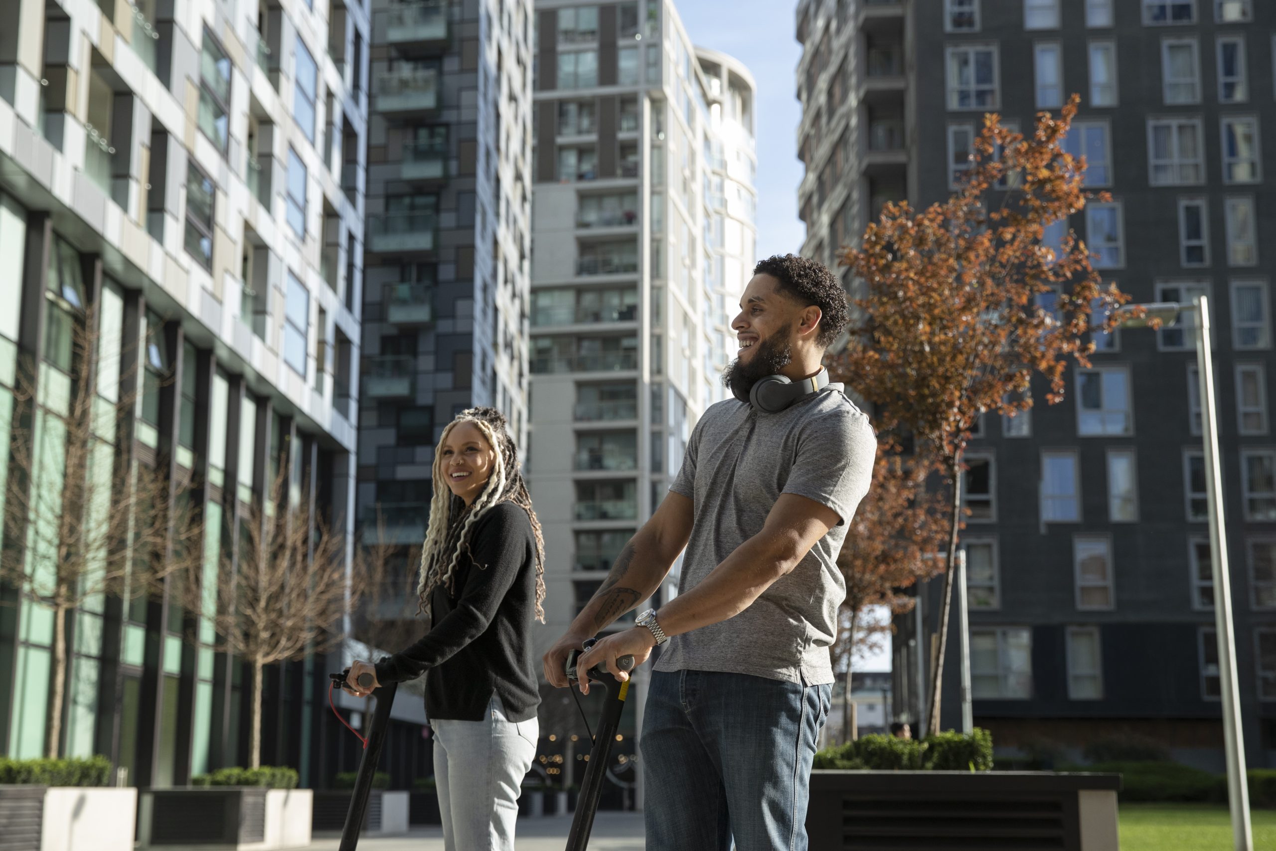 Um homem e uma mulher sorridentes andam de patinete elétrico em uma área urbana moderna, cercada por prédios de apartamentos altos e modernos sob a luz do sol.
