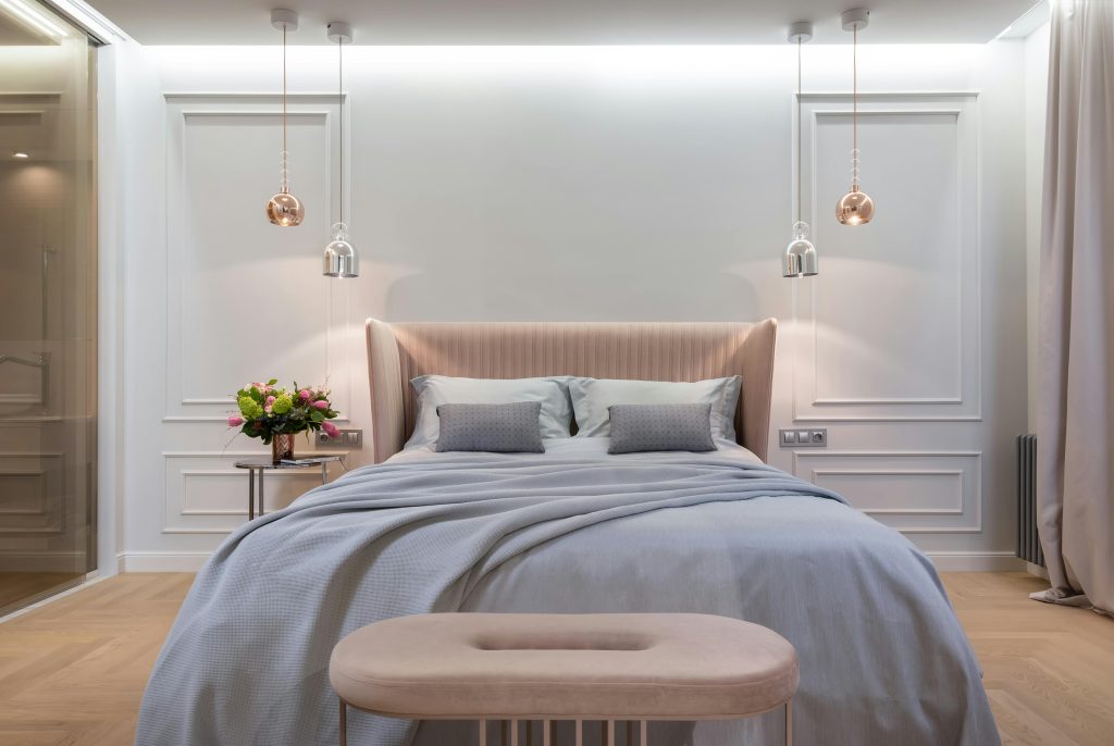 Quarto moderno com cama de casal queen size, cabeceira estofada rosa claro e luminárias pendentes de cristal sobre mesinhas de cabeceira em um ambiente com paredes brancas decoradas com molduras.