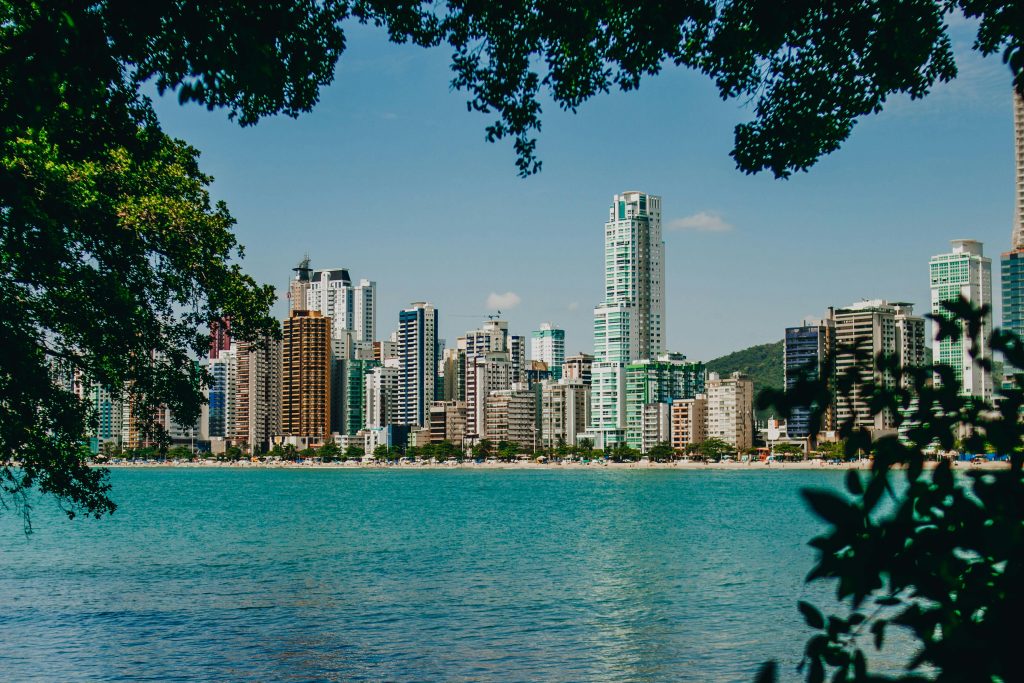 ista da Praia Central de Balneário Camboriú através da folhagem de árvores, mostrando o mar azul em primeiro plano e o horizonte repleto de grandes arranha-céus sob um céu claro.