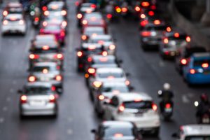 Trânsito intenso em avenida urbana com carros e motos parados, visto de forma desfocada ao entardecer.
