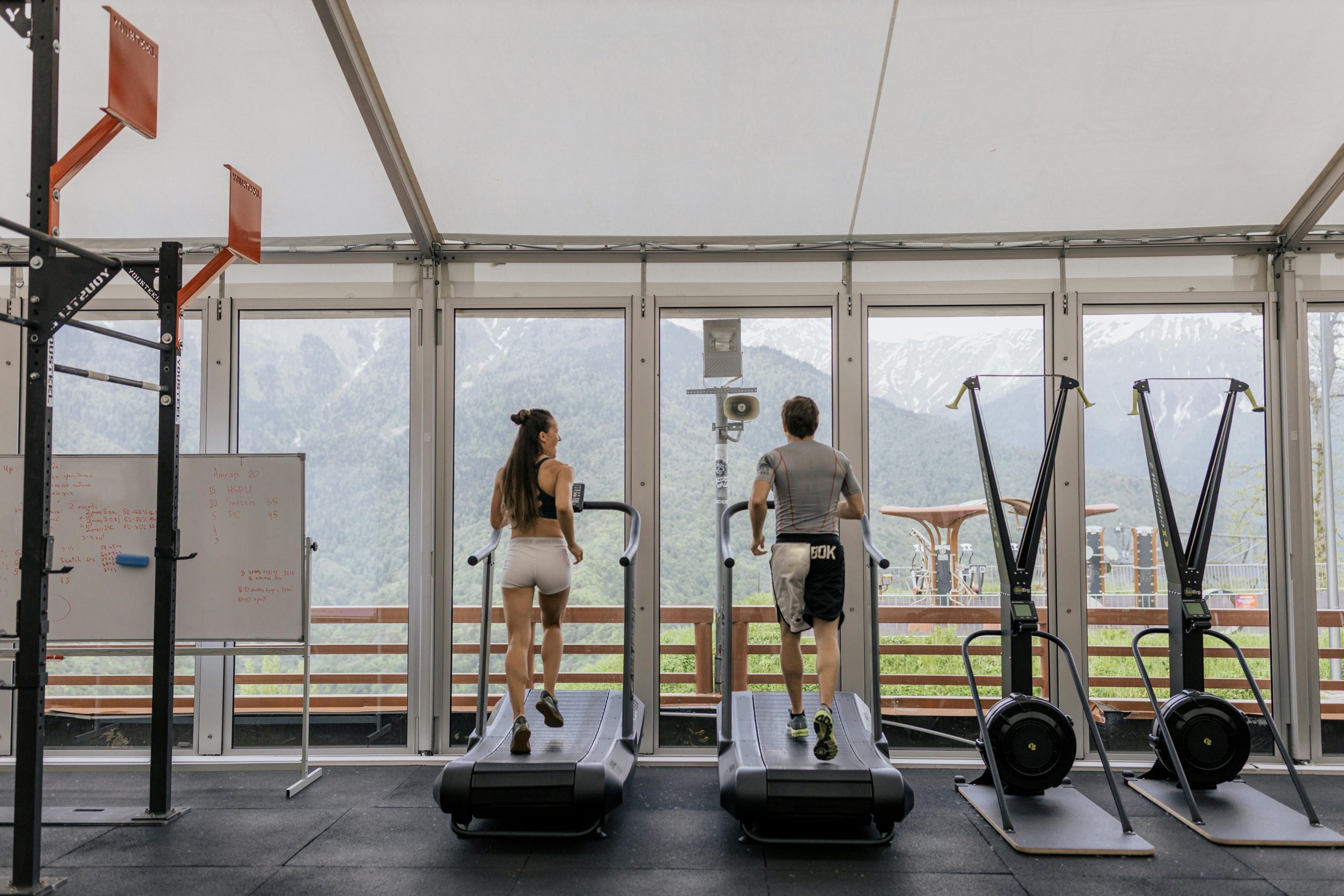 Duas pessoas correndo em esteiras em uma academia com vista para montanhas através de grandes janelas.